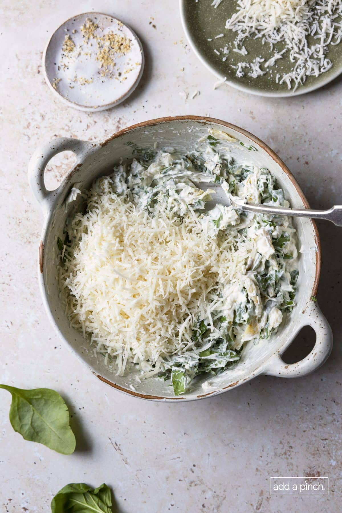 Grated parmesan cheese being stirred into spinach artichoke dip recipe in a white baking dish.