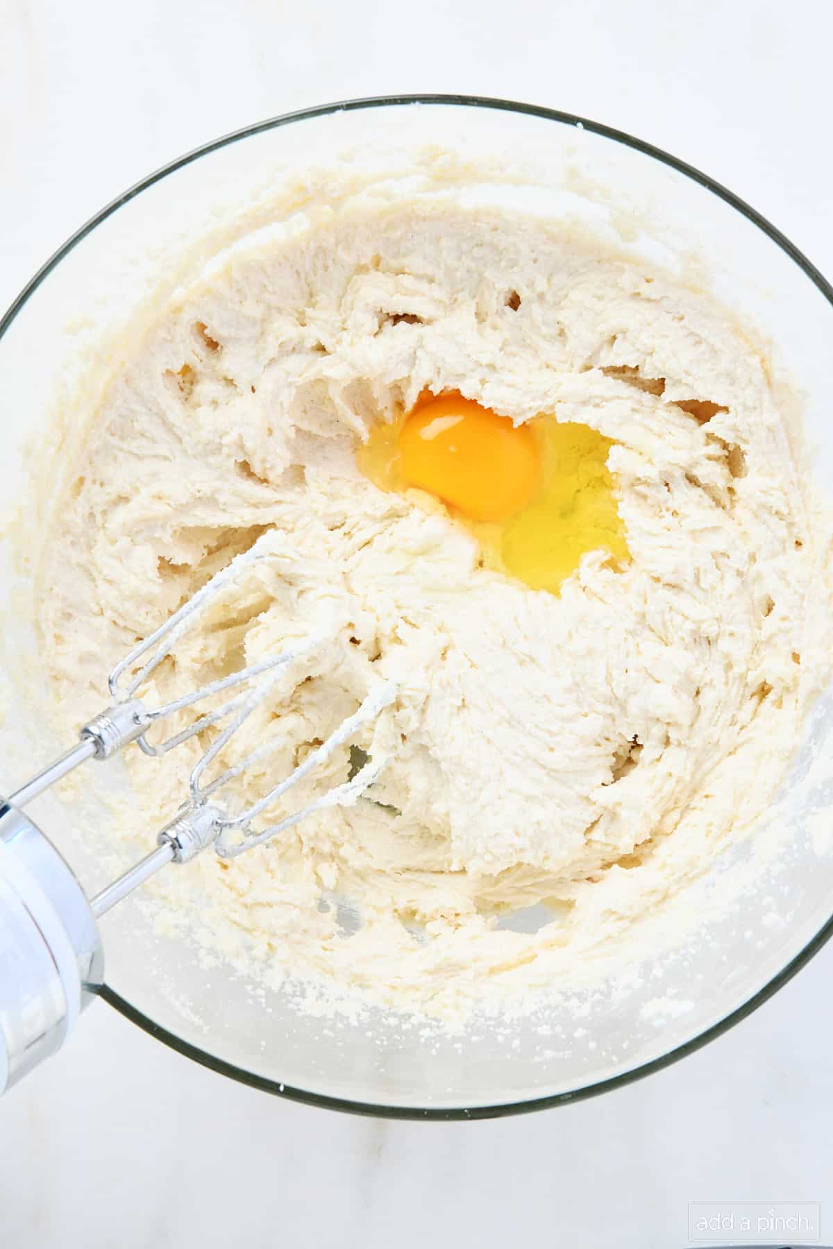 In a glass mixing bowl, an egg is being incorporated into the creamed butter, sugar, shortening mixture with the hand mixer. 