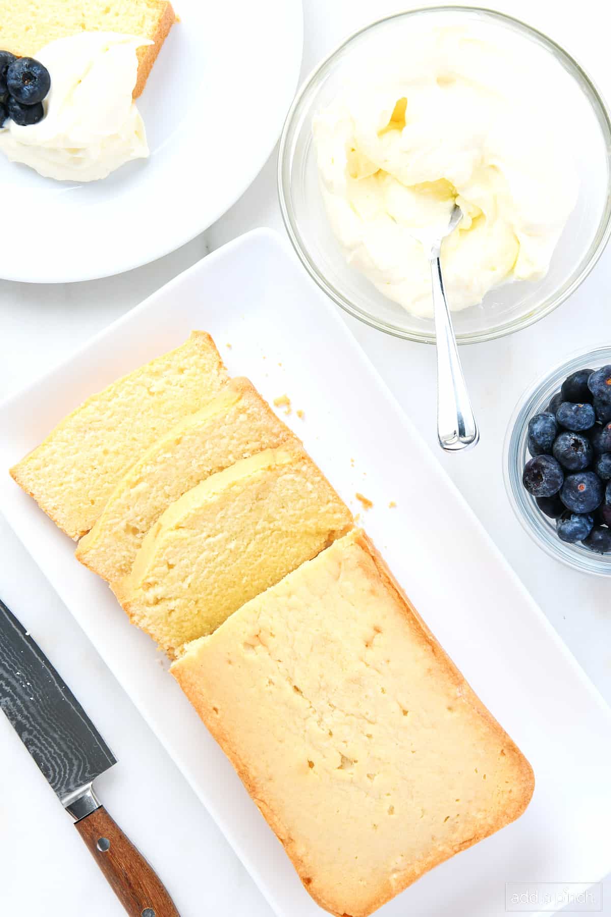Overhead shot of loaf size pound cake that has been sliced, along with bowl of homemade whipped cream and fresh blueberries. A slice is served on a white plate and a knife is nearby.
