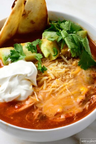 Bowl of chicken tortilla soup garnished with tortilla strips, avocados, sour cream, and shredded cheese, served in a white bowl.