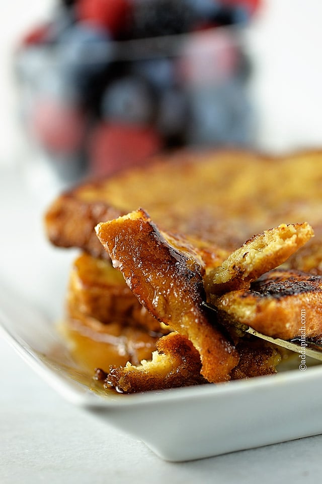 Bite of French toast on a fork on a white plate with more french toast and berries in the background.