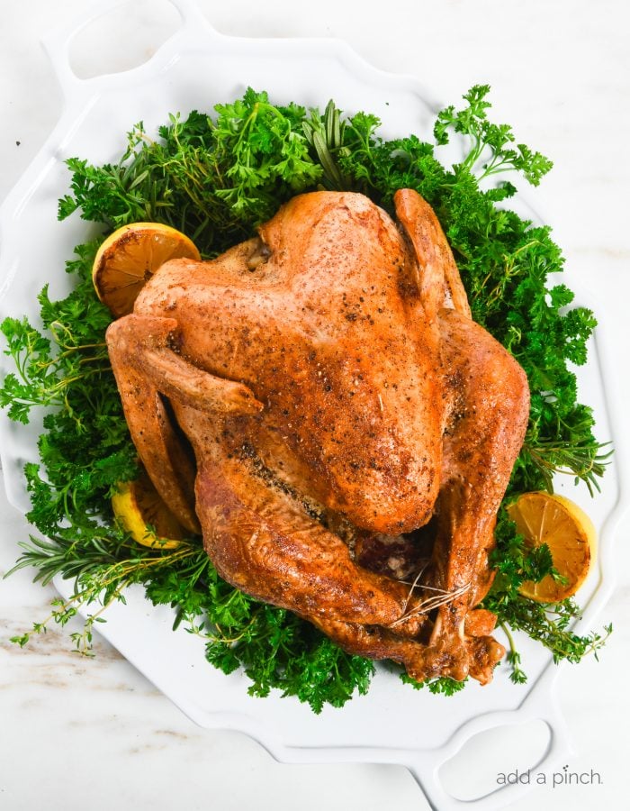 Overhead photo of a golden brown turkey on a large white platter garnished with fresh herbs, lemon, salt and pepper.