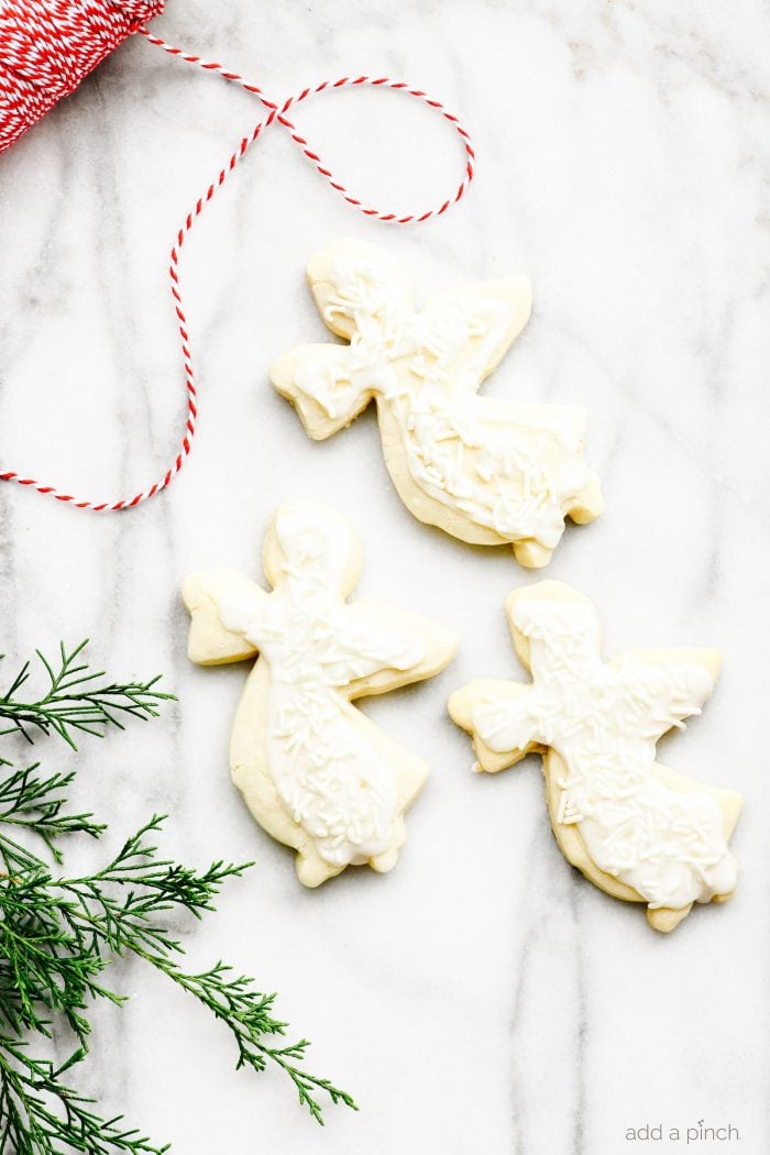 Cookies in shape of angels decorated with white icing and white sprinkles. Surrounded by greenery and red and white twine on a marble board. 